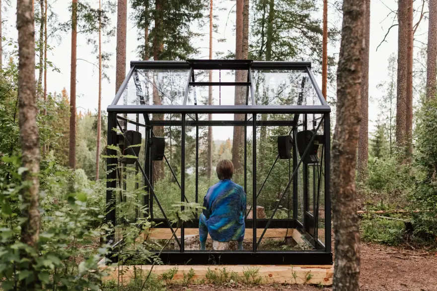 kuvassa henkilö istuu taideteoksen (lasisen huoneen) sisällä luonnossa