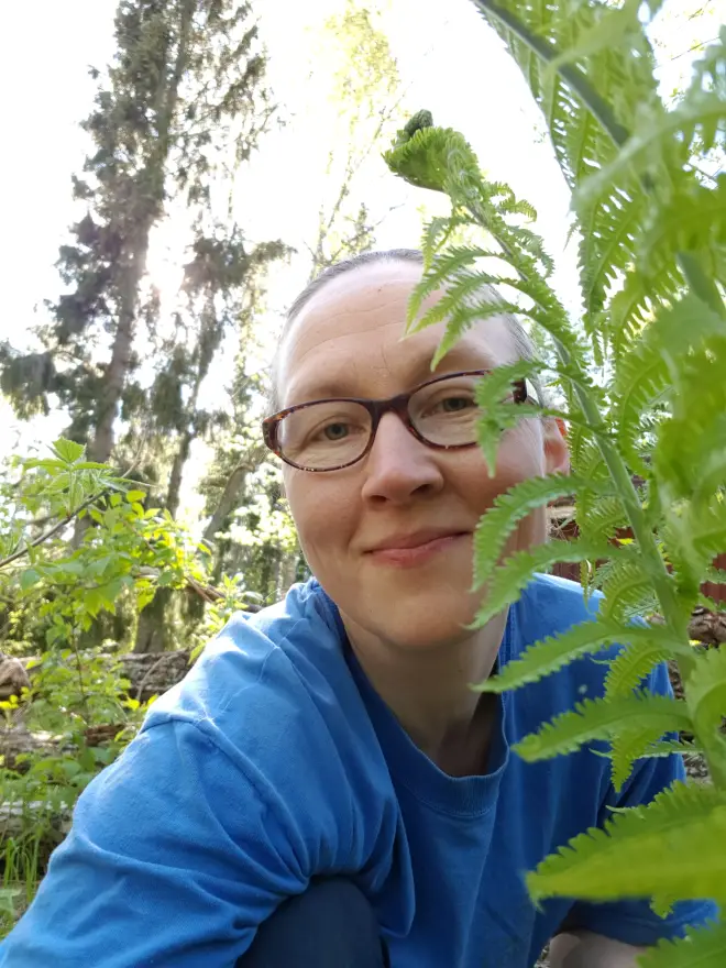 Saara Korpela's selfie in a forest behind a fern leaf. Saara has a blue shirt.